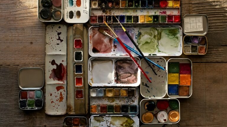 Well-used tins of watercolour paint and brushes on a worn wooden table.